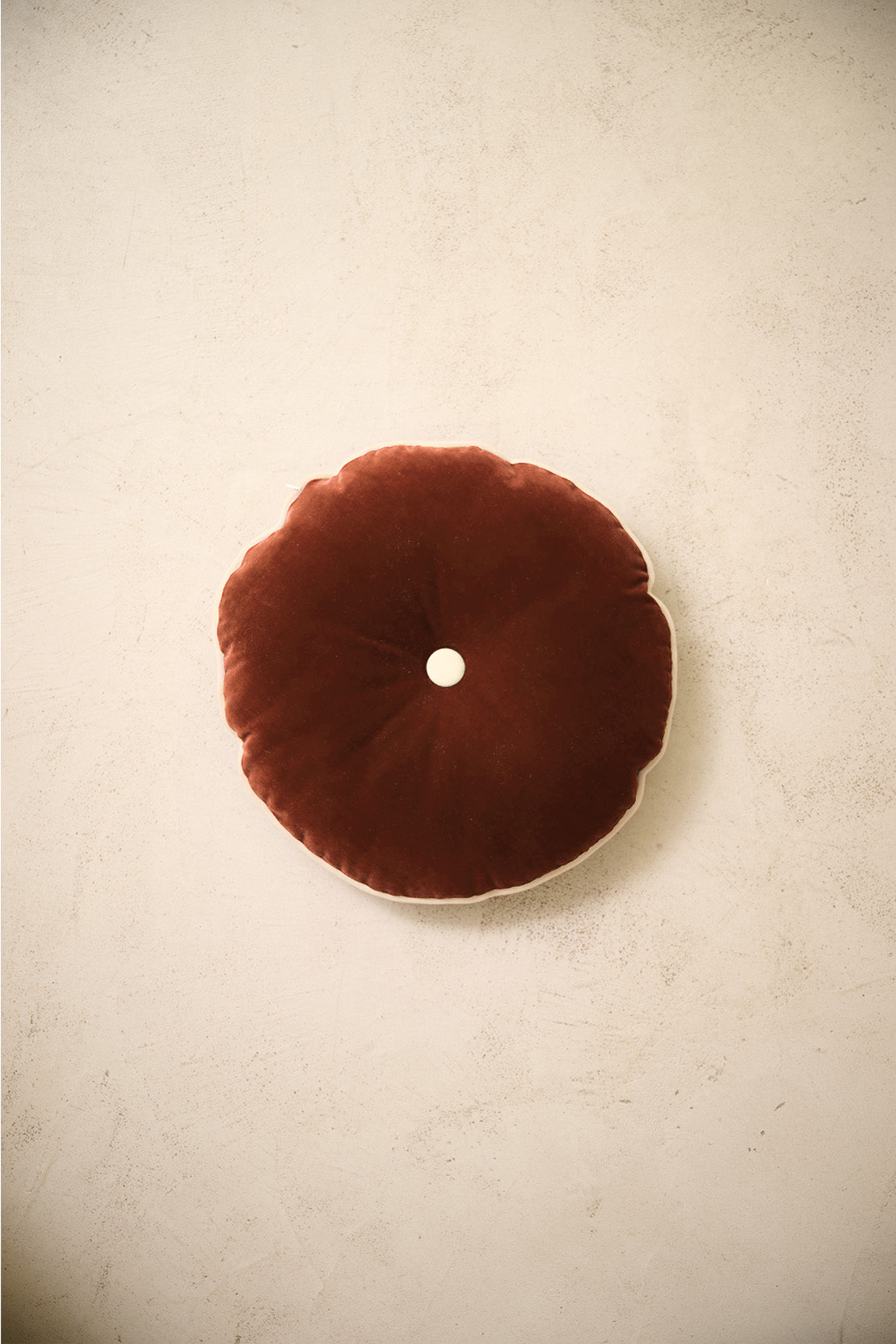 coussin rond MOON en velours cognac à bouton écru, posé sur un mur en enduit crème