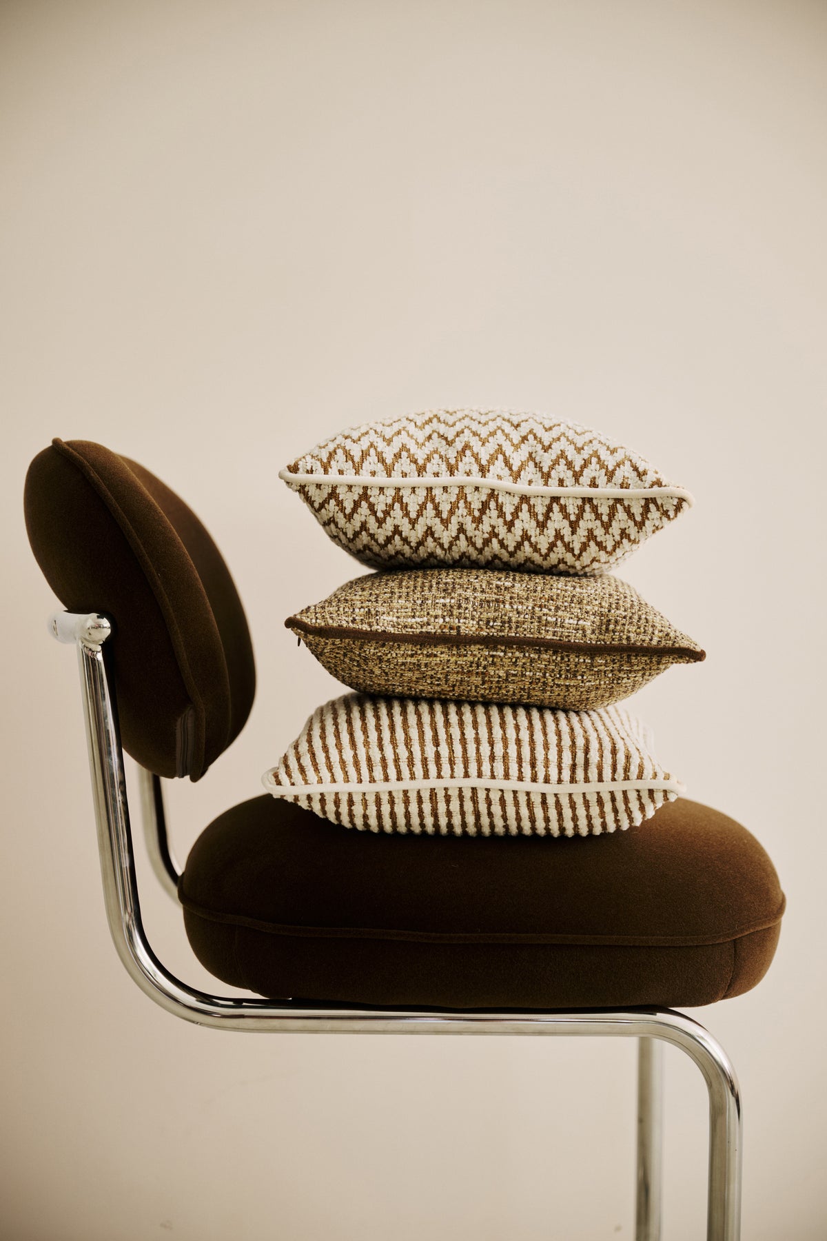 trois coussins LODGE à motifs écru et marron empilés sur une chaise en velours brun