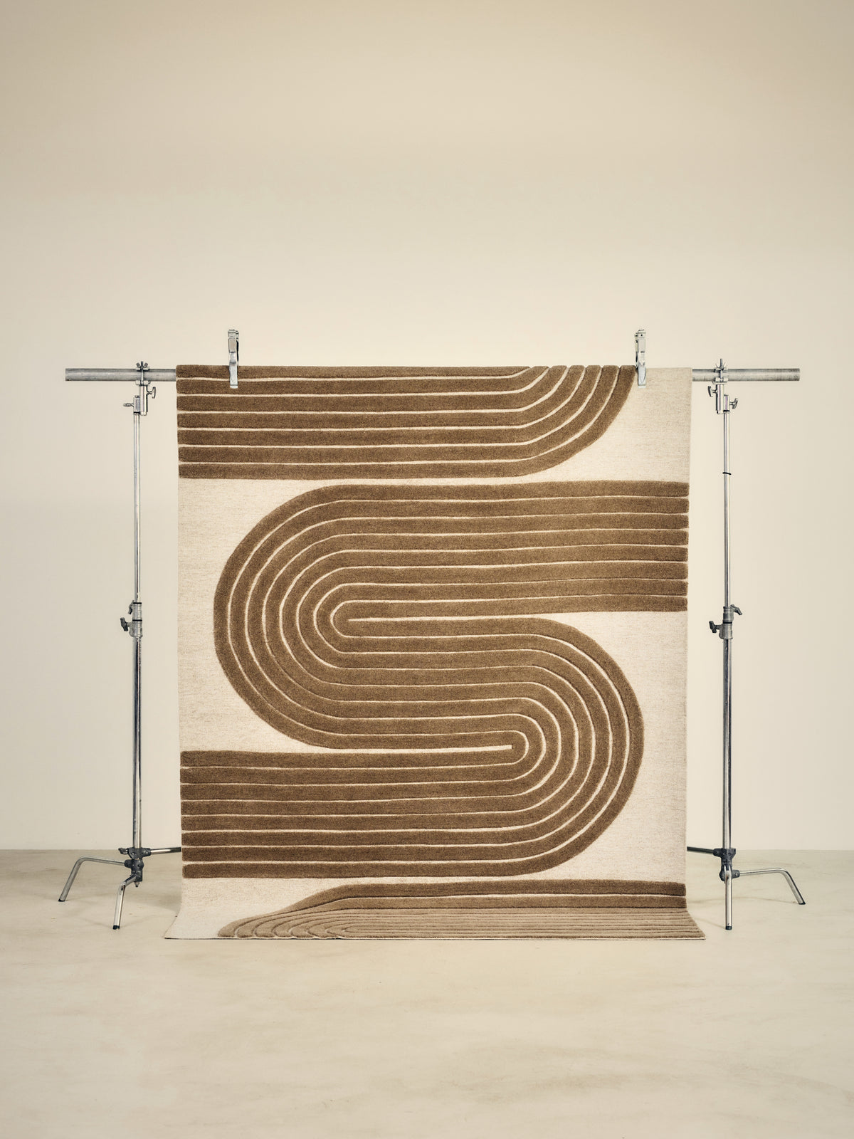 tapis ARCO beige à larges courbes brunes en relief, suspendu sur un portant de studio