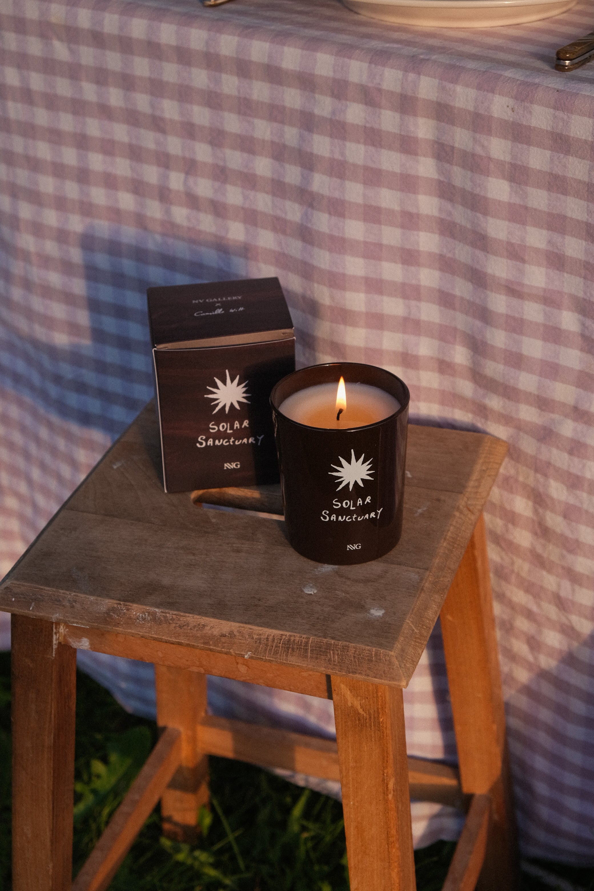 bougie Solar Sanctuary NVG x Wiwoos - verre ambré allumé et coffret - mise en situation tabouret jardin vichy