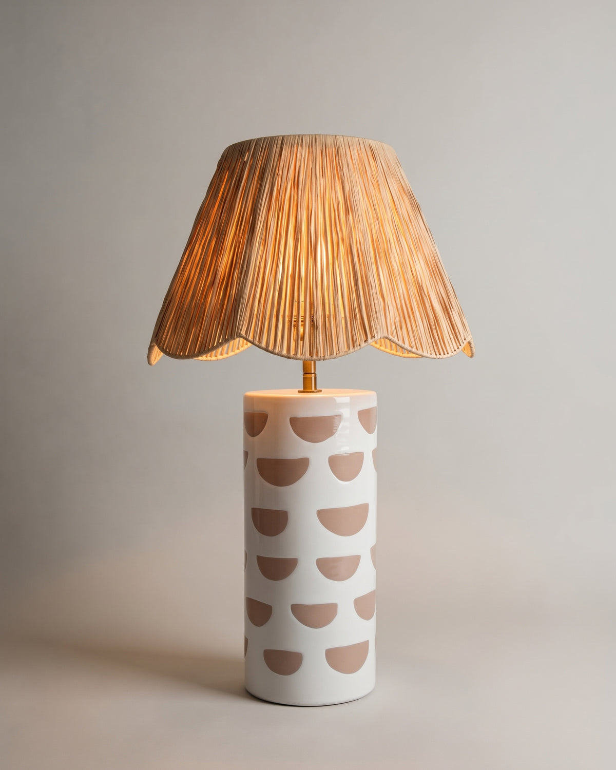 lampe de table MARA allumée, céramique blanche à demi-lunes rosées et raphia festonné